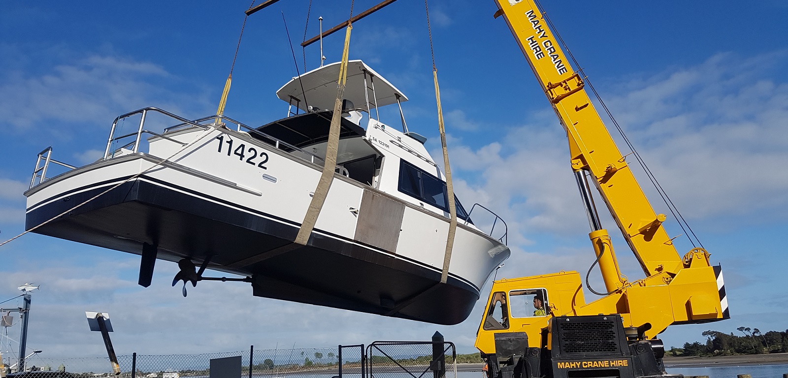 Mahy Crane Hire mobile crane lifting a large boat with slings at a waterfront site